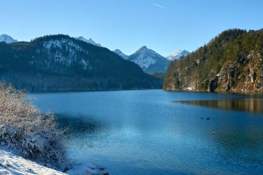 Bu yüksek çözünürlüklü fotoğraf sakin bir kış manzarasını yakalar. Sahne, etrafındaki dağları ve gökyüzünü yansıtan kristal berrak, derin mavi suya sahip büyük, sakin bir göle sahiptir. Göl karla kaplı, yemyeşil ormanlarla çevrili.