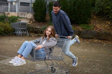 Canlı genç bir çift sıradan bir alışveriş arabasını bu büyüleyici sonbahar sahnesinde spontane bir neşe ve romantik macera sembolüne dönüştürür. Kareli fanila gömlekli ve açık mavi kotlu bir kadın neşe içinde oturuyordu.