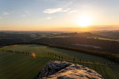 Göz kamaştırıcı günbatımı, Lilienstein 'in bakış açısından görüldüğü gibi yemyeşil tarlalar ve yuvarlanan tepeler üzerine altın bir ışık saçar. Elbe Kum Taşı Dağları dramatik bir zemin oluşturur.