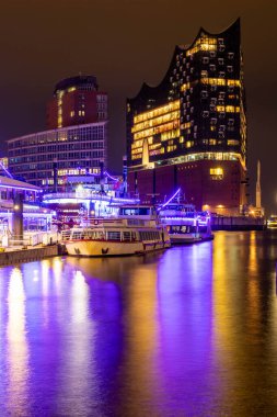 Canlı ışıklar geceleri Hamburg 'daki rıhtım boyunca tekneleri aydınlatıyor. Uzun süre maruz kalınca, sakin sulardaki güzel yansımalar şehrin büyüleyici atmosferini güçlendiriyor..