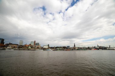 Panoramik bir kıyı manzarası Hamburg 'un etkileyici ufuk çizgisini çeşitli binalar ve canlı limanlarla yakalar. Tekneler ve gemiler dramatik bir gökyüzünün altında.
