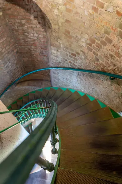 Yuvarlak çelik bir merdiven, taş duvarlar ve yeşil korkulukların bulunduğu eski tuğla binanın içine doğru iniyor. Dokular ve renkler tarihi bir atmosfer yaratıyor, keşifleri davet ediyor..