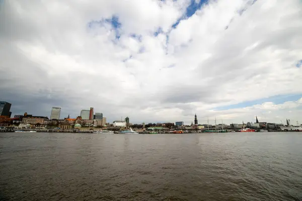 Panoramik bir kıyı manzarası Hamburg 'un etkileyici ufuk çizgisini çeşitli binalar ve canlı limanlarla yakalar. Tekneler ve gemiler dramatik bir gökyüzünün altında.