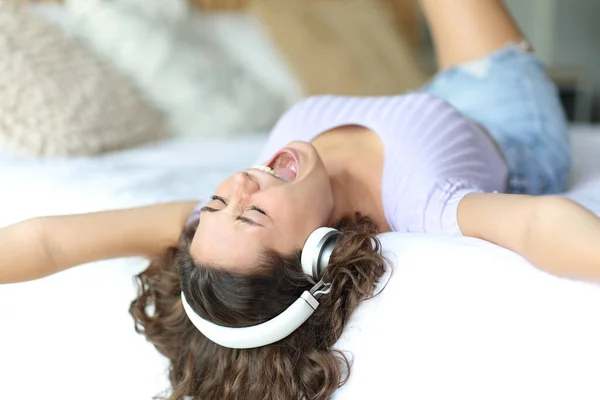 一个兴奋的女人 头戴耳机 躺在房间的床上 一边唱歌一边听音乐 — 图库照片
