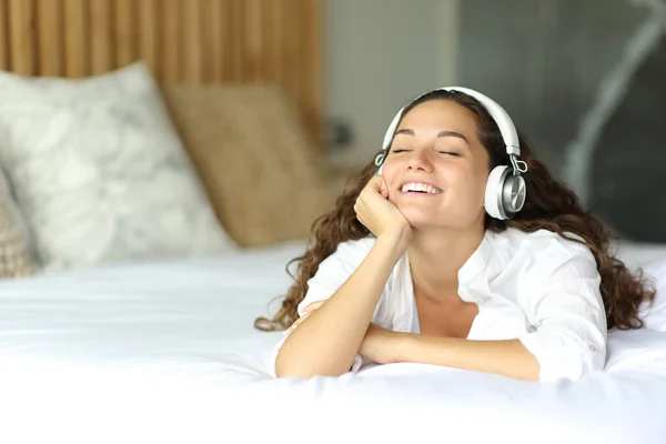躺在床上听音乐的快乐女人卧室里有耳机 — 图库照片