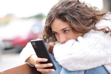 Üzgün kadın caddedeki bankta oturan akıllı telefonu kontrol ediyor.