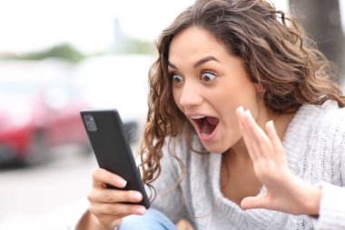 Şaşkın kadın sokakta akıllı telefon içeriğini kontrol ediyor.