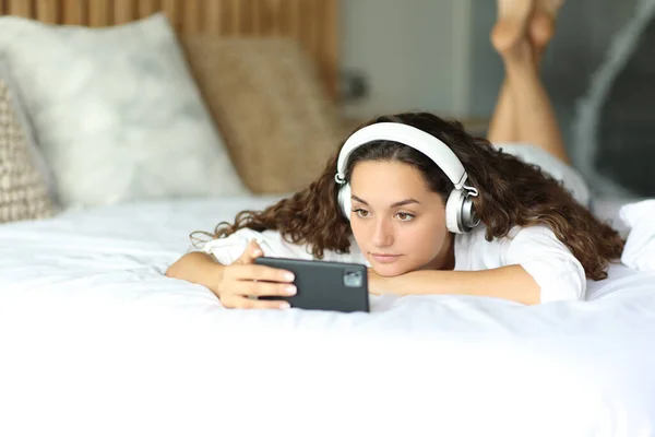 Serious woman watching videos on phone lying on a bed