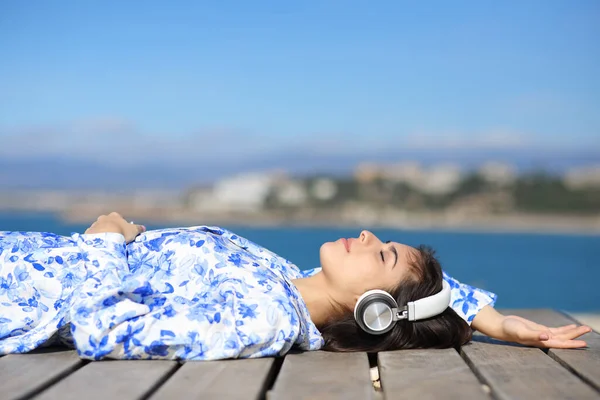 Kobieta Relaksująca Się Słuchając Muzyki Słuchawką Leżącą Drewnianej Platformie Plaży — Zdjęcie stockowe