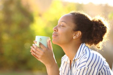 Kafası rahat, kahve içen ve parktaki aromayı koklayan siyah bir kadının profili.