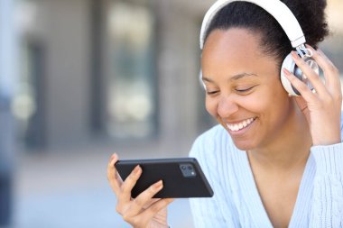 Kulaklık takan mutlu siyah kadın sokakta yatay akıllı telefondan video izliyor.