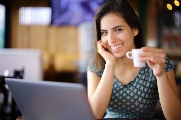 Happy woman with laptop drinking in a coffee shop looking at camera