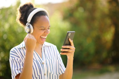 Heyecanlı siyah kadın kulaklık takıyor ve parkta telefonla müzik dinliyor.