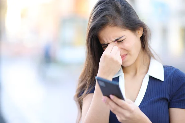 Stressed woman suffering eyestrein holding phone in the street