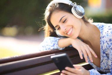 Parkta bankta oturmuş telefon ve kulaklıkla müzik dinleyen mutlu bir kadın.