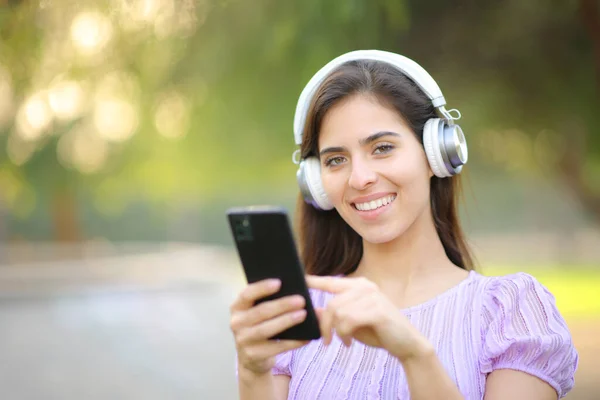 ハッピーな女性はヘッドフォンを着て 公園でカメラを見ている音楽を聴く携帯電話を持っています — ストック写真