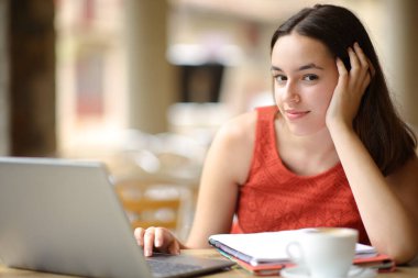 Kendine güvenen bir öğrenci bar terasında dizüstü bilgisayarla sana bakıyor.