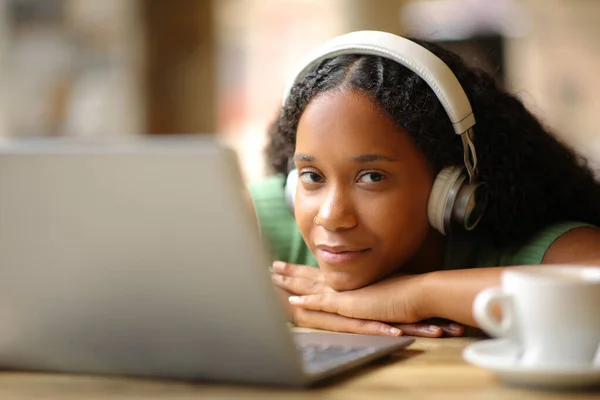 Black woman with headphone and laptop looks at you in a coffee shop terrace