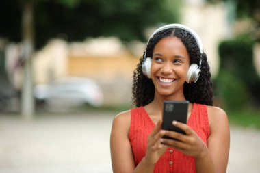 Kulaklık takmış, elinde telefonla müzik dinleyen mutlu siyahi bir kadının portresi.