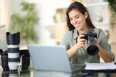 Mutlu fotoğrafçı evde elinde kamerayla bilgisayarı kontrol ediyor.