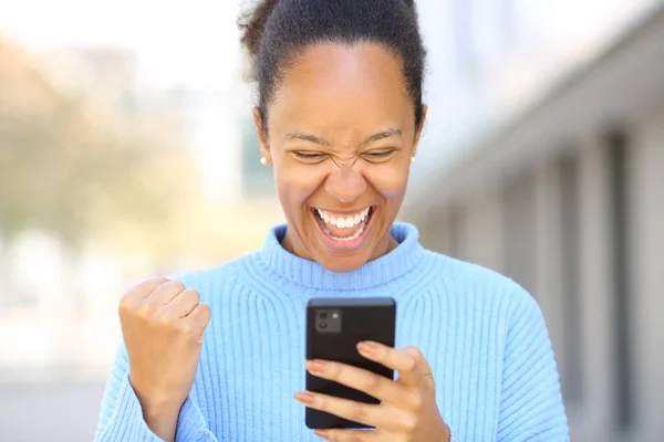 Front view portrait of an excited black woman checking good news on phone in the street