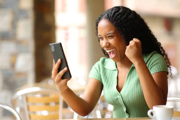 Excited black woman checking smart phone in a bar terrace