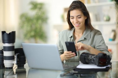Bilgisayarlı ve kameralı mutlu fotoğrafçı evde telefonu kontrol ediyor.