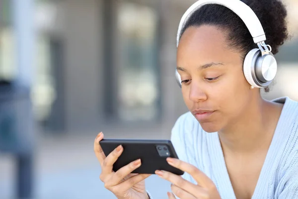 Black woman with headphone watching video online on phone in the street