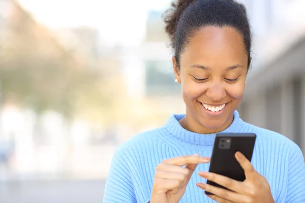 Front view portrait of a happy black woman using cell phone in the street with copy space