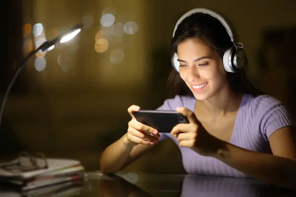 自宅で携帯電話でビデオを見ている夜にヘッドフォンを身に着けている幸せな女性 — ストック写真