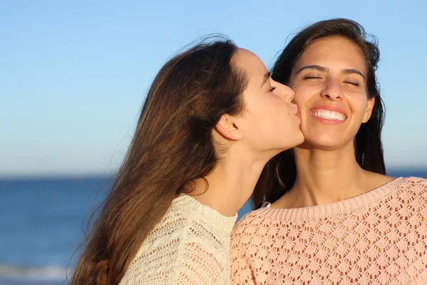 Happy woman kissing a friend on cheek on the beach at sunset