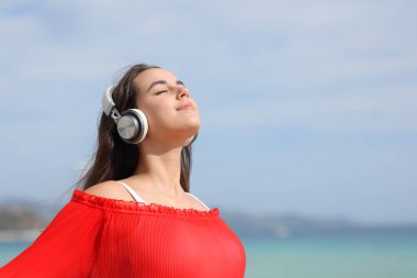 Kırmızı elbiseli rahat bir kadın temiz hava soluyor. Plajda kulaklıkla müzik rehberi dinliyor.