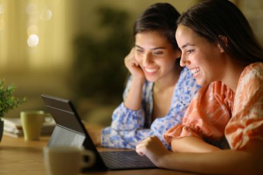 Two happy friends in the night watching videos on tablet at home clipart