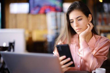 Bilgisayarlı ciddi bir kadın barın içinde cep telefonu içeriğini kontrol ediyor.