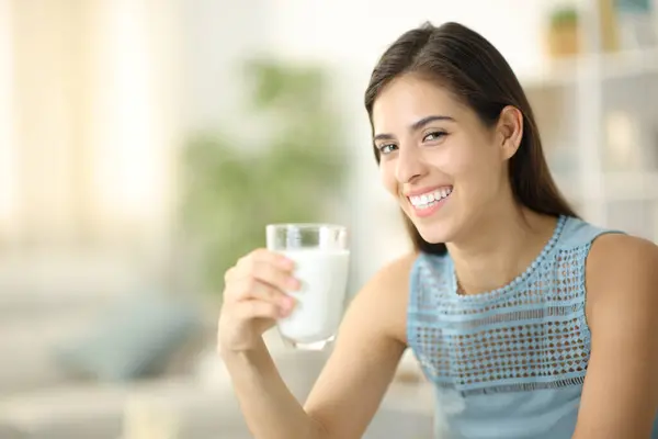 自宅でミルクのグラスを握っているカメラで笑顔の幸せな女性 — ストック写真