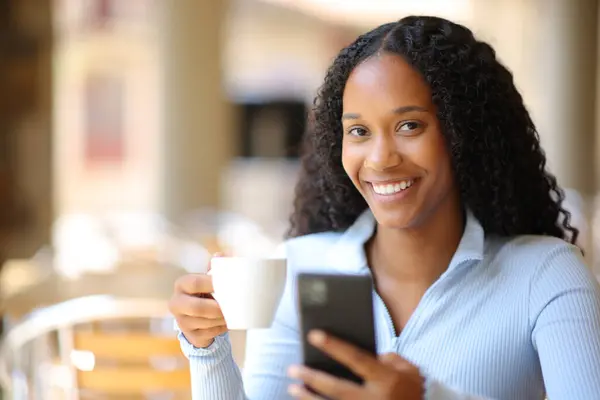 Happy black woman holding phone and coffee cup in a restaurant terrace
