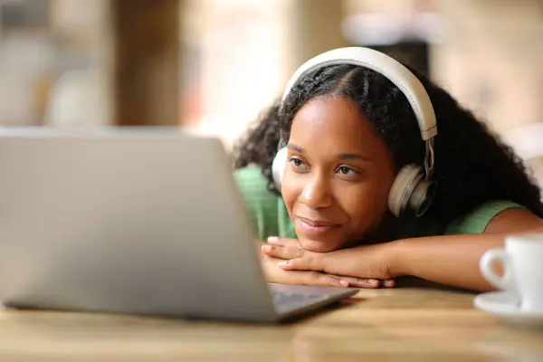 满意的黑人妇女在餐馆的阳台上通过耳机观看笔记本电脑上的媒体内容 图库图片