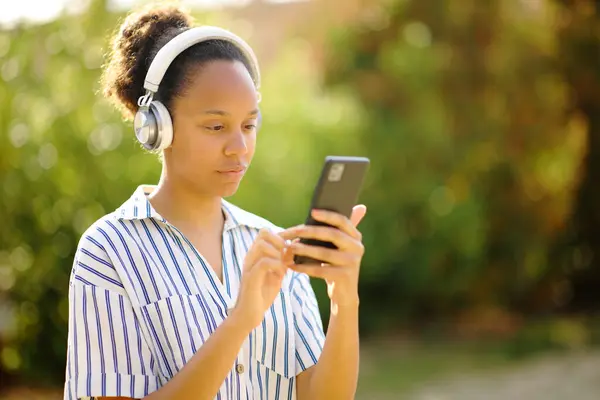 在花园里用电话走来走去 戴耳机的黑人妇女 免版税图库照片