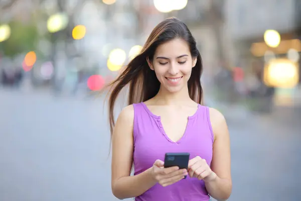 一个快乐的女人在街上用智能手机走路的正面画像 图库图片
