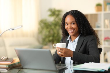 Evde elinde kahve fincanıyla kameraya bakarak poz veren mutlu televizyon çalışanı.
