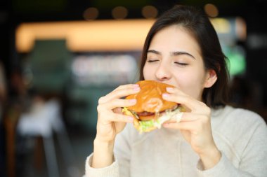 Lokantanın içinde hamburger yiyen mutlu bir kadın.