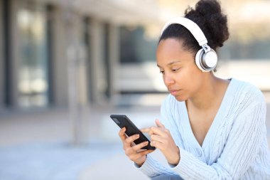 Kulaklık takan siyah bir kadın sokakta telefon kontrol ediyor.