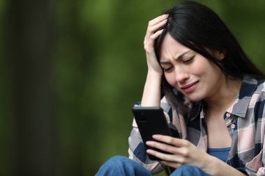 Endişeli Asyalı kadın cep telefonundan kötü haberleri kontrol ediyor ve parkta şikayet ediyor.