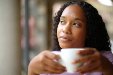 Düşünceli, ciddi, siyah bir kadın bir bar terasında kahve içiyor.