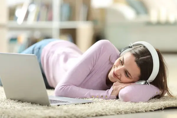 stock image Happy woman wearing headphone watching media on laptop lying on a carpet at home