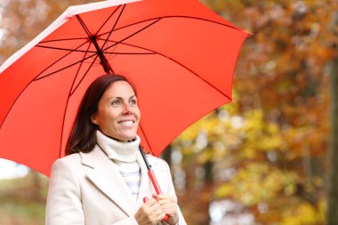 Kırmızı şemsiye altında mutlu, orta yaşlı bir kadın sonbahar ormanlarını seyrediyor.