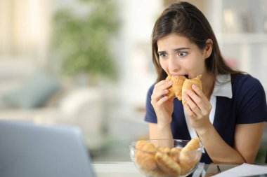 Bilgisayarda medyayı izleyen bir kadın evde zorla kruvasan yiyor.