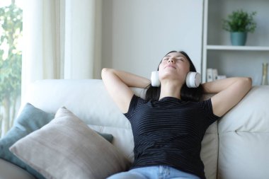 Asyalı kadın evde bir kanepede oturmuş kulaklıkla müzik dinliyor.