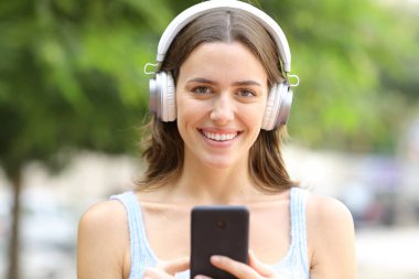 Parkta elinde akıllı bir telefonla kulaklık takan mutlu bir kadının portresi.