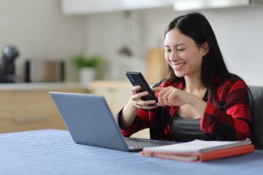 Asyalı öğrenci evdeki mutfakta bilgisayar ve telefon kullanıyor.